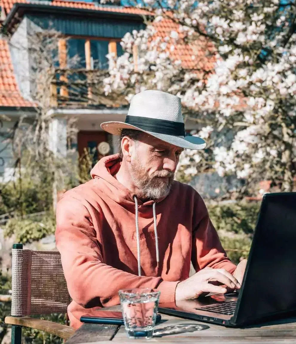 Arwed Grön, Gründer von GROENIE.AI, mit Hut und orangem Hoodie am Laptop im Garten